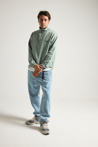 Personne portant un sweat demi-zip Sauce Pigment vert argenté de coupe décontractée, associé à un pantalon bleu clair et des baskets, posant sur un fond neutre. | TP_IMAGE_TYPE=MODEL_FRONT