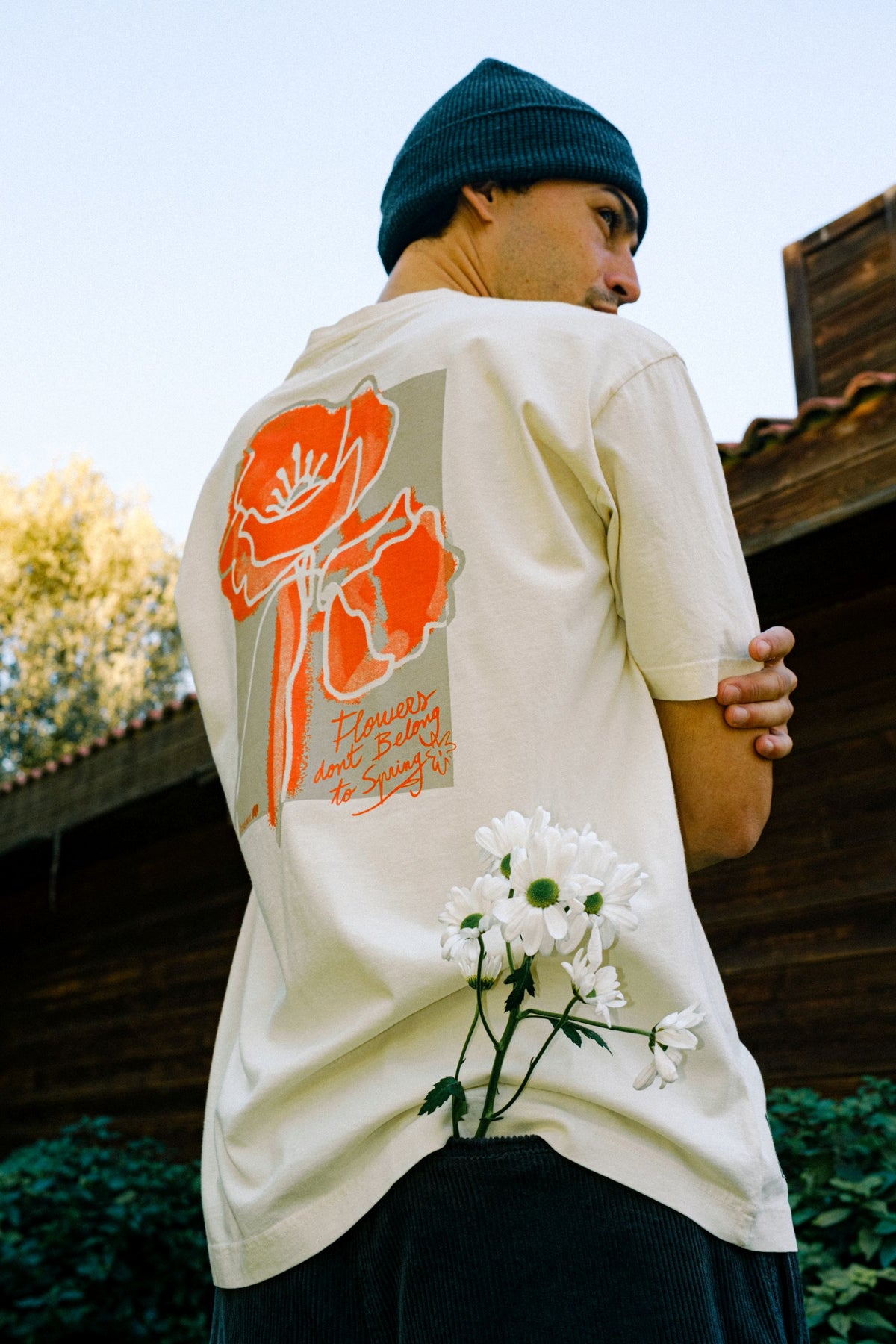 Vista posteriore di una persona che indossa una t-shirt grafica viola e un berretto, in piedi su un sentiero coperto di foglie mentre tiene uno skateboard. TP_IMAGE_TYPE=LIFESTYLE