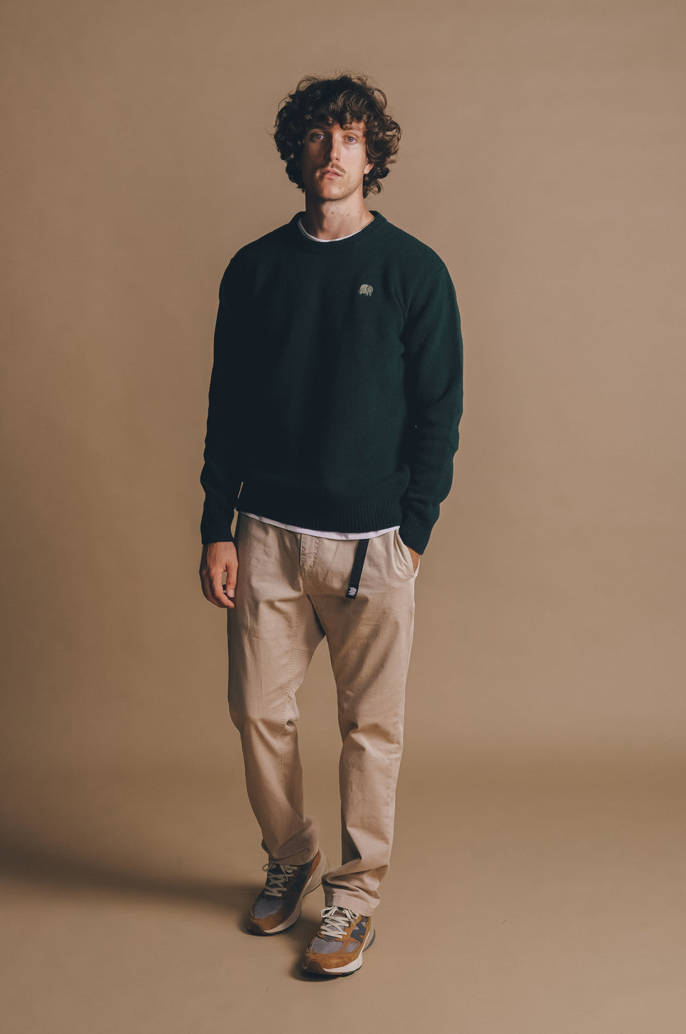 Persona che indossa un maglione in maglia verde pino abbinato a pantaloni beige chiaro e sneakers, in piedi su uno sfondo neutro. | TP_IMAGE_TYPE=MODEL_FRONT