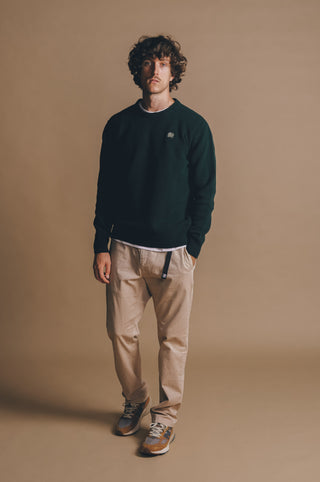 Persona che indossa un maglione in maglia verde pino abbinato a pantaloni beige chiaro e sneakers, in piedi su uno sfondo neutro. | TP_IMAGE_TYPE=MODEL_FRONT