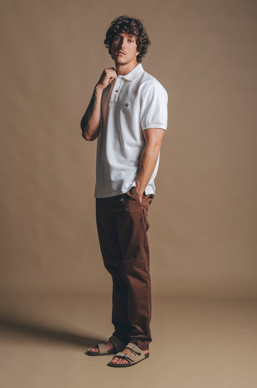 Persona che indossa la Polo Shirt Organica Tinta a Pigmento Alcalali in Almond Milk, abbinata a pantaloni marroni e sandali, in posa pensierosa su uno sfondo neutro | TP_IMAGE_TYPE=MODEL_FRONT