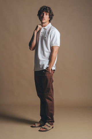 Persona che indossa la Polo Shirt Organica Tinta a Pigmento Alcalali in Almond Milk, abbinata a pantaloni marroni e sandali, in posa pensierosa su uno sfondo neutro | TP_IMAGE_TYPE=MODEL_FRONT