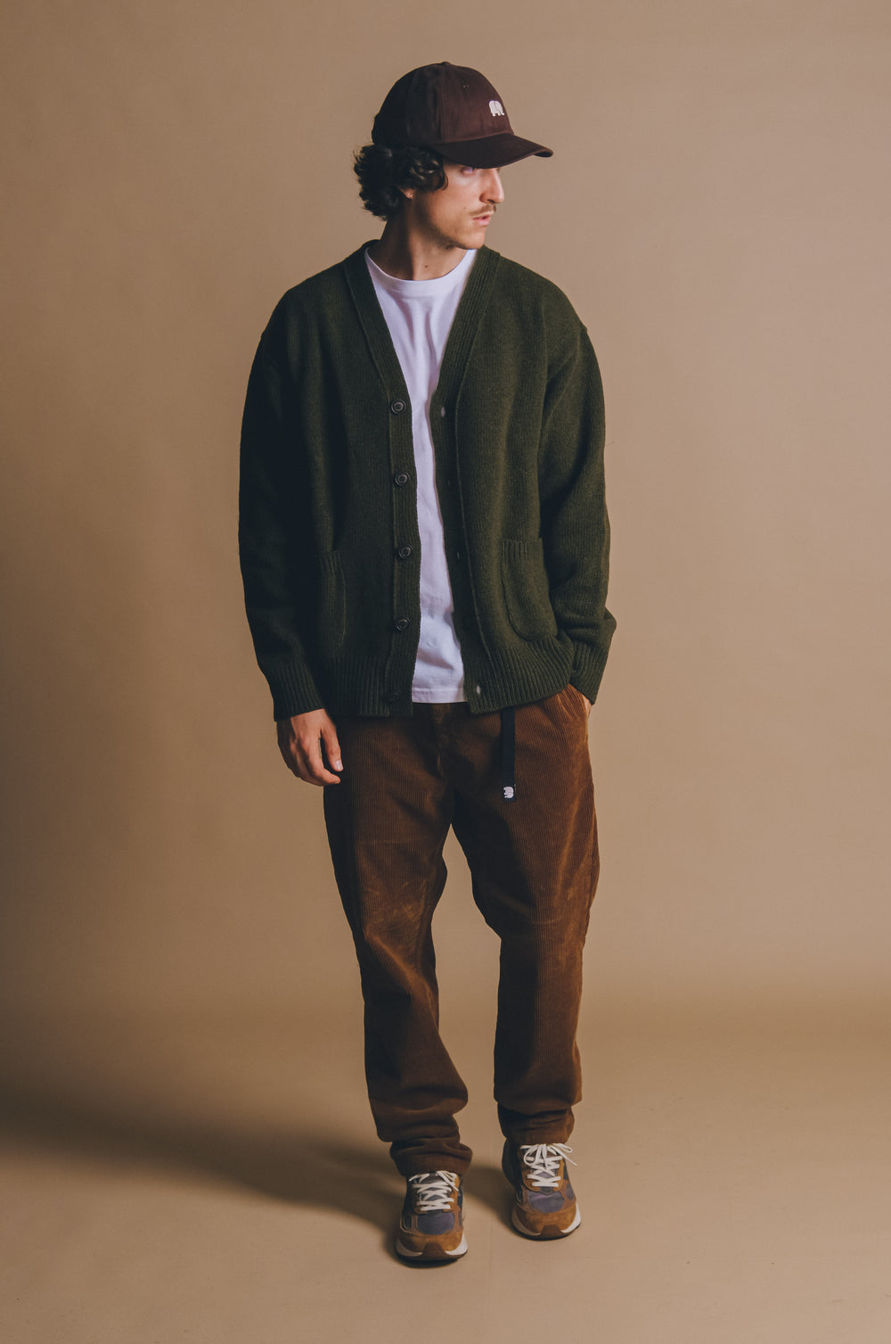 Persona che indossa un Relleu Cardigan in Ivy Green, abbinato a una camicia bianca e a pantaloni marroni, in posa su uno sfondo neutro. | TP_IMAGE_TYPE=MODEL_FRONT