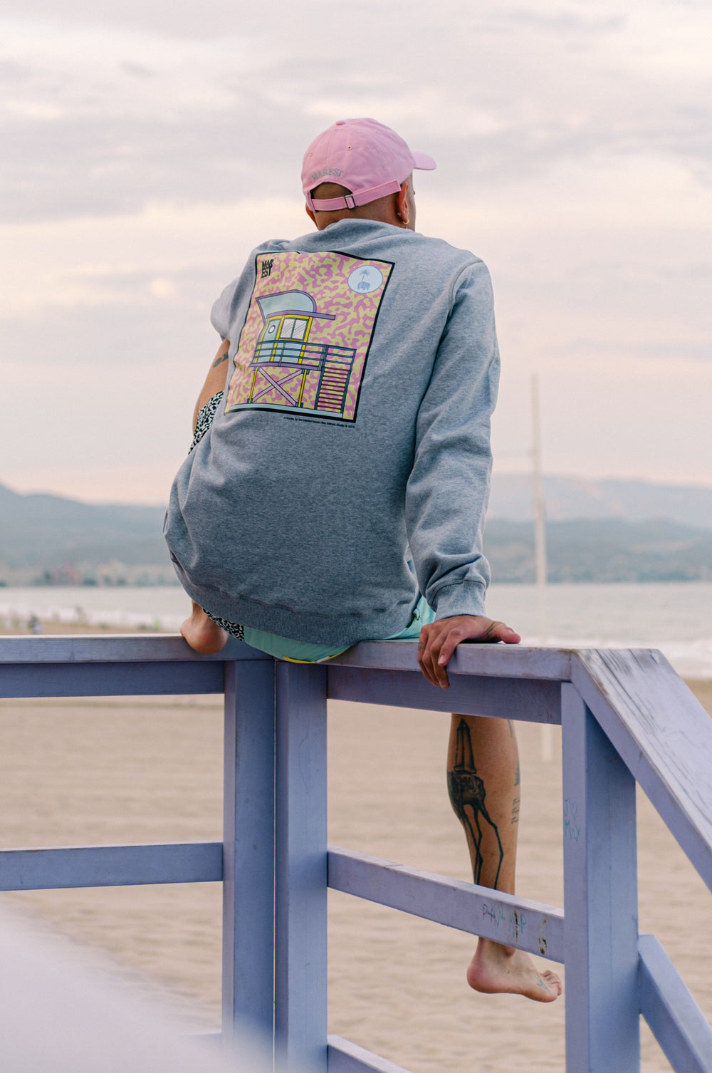 Persona che indossa una felpa Antonyo Marest x Trendsplant Art Hut in Sport Grey, seduta su una ringhiera vicino alla spiaggia | TP_IMAGE_TYPE=MODEL_BACK
