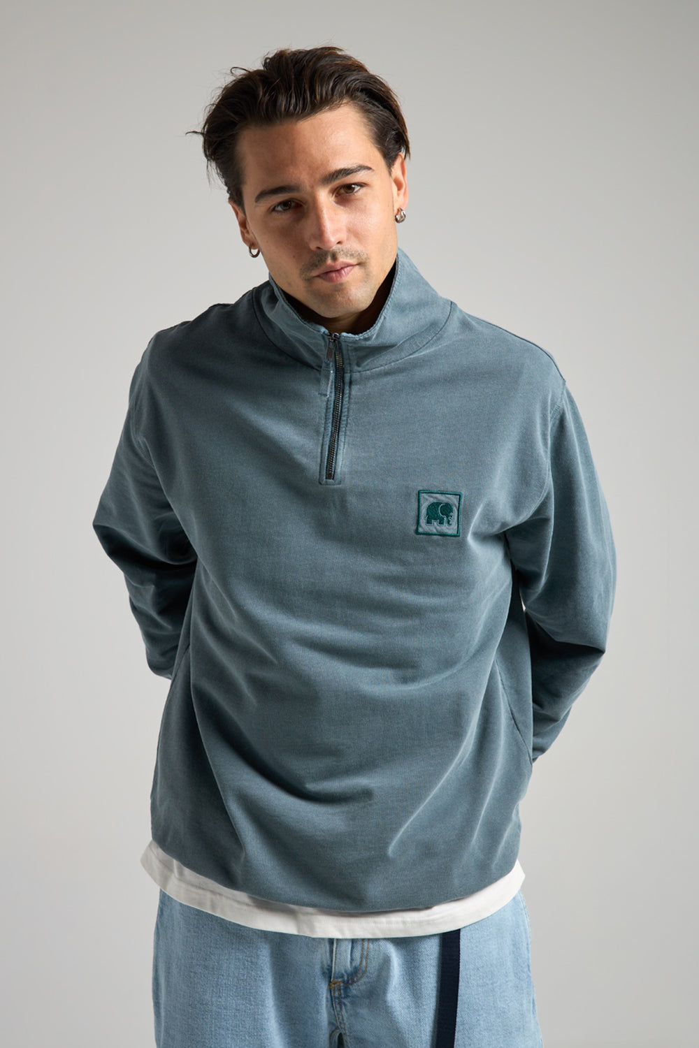 Persona che indossa Sauce Pigment Dyed Quarter Zip Sweatshirt in Elm Green, con chiusura a mezza zip e una patch con il logo sul petto | TP_IMAGE_TYPE=MODEL_FRONT