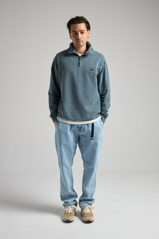 Persona che indossa una Sauce Pigment Dyed Quarter Zip Sweatshirt in Elm Green, abbinata a pantaloni azzurro chiaro e sneakers, in posa su uno sfondo neutro. | TP_IMAGE_TYPE=MODEL_FRONT
