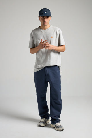 Persona che indossa una t-shirt organica Essentials in grigio mélange, abbinata a pantaloni blu navy e sneakers casual, in posa in uno studio fotografico | TP_IMAGE_TYPE=MODEL_FRONT