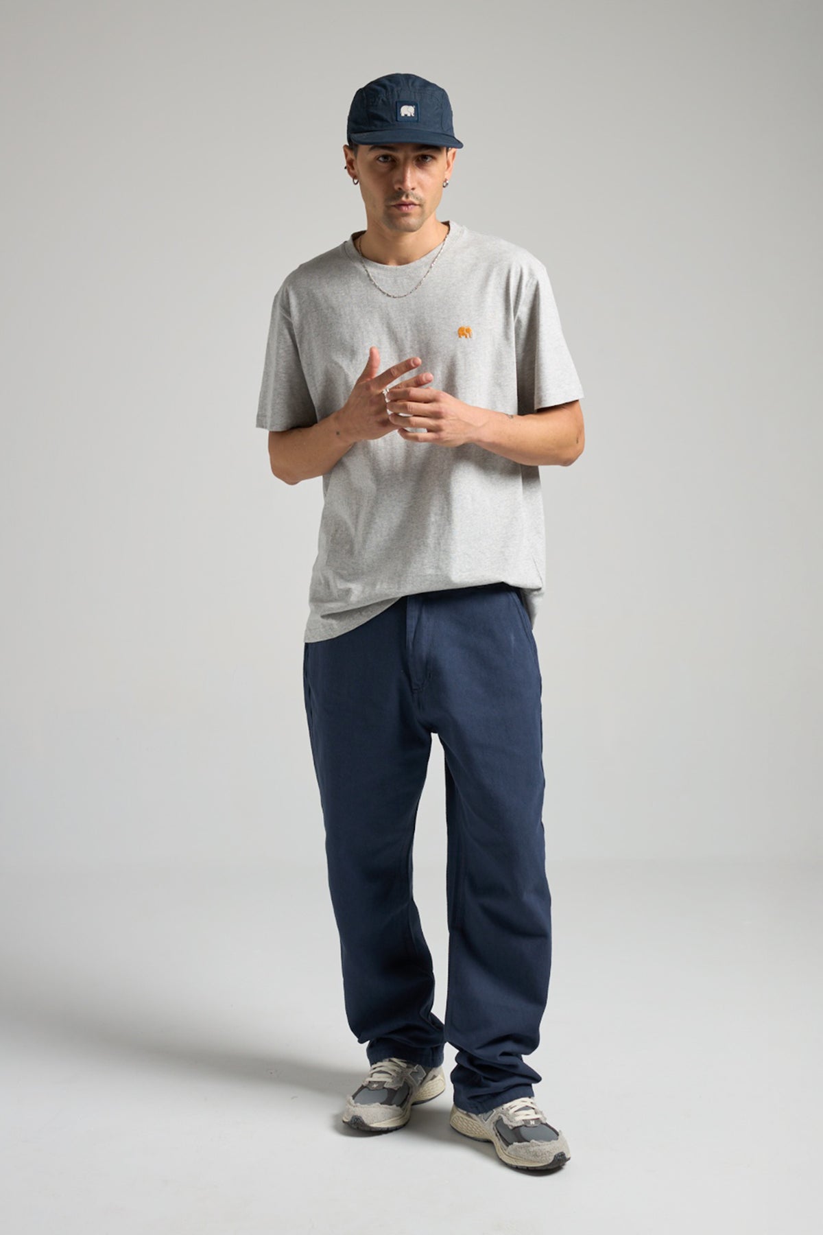 Persona che indossa una t-shirt organica Essentials in grigio mélange, abbinata a pantaloni blu navy e sneakers casual, in posa in uno studio fotografico | TP_IMAGE_TYPE=MODEL_FRONT