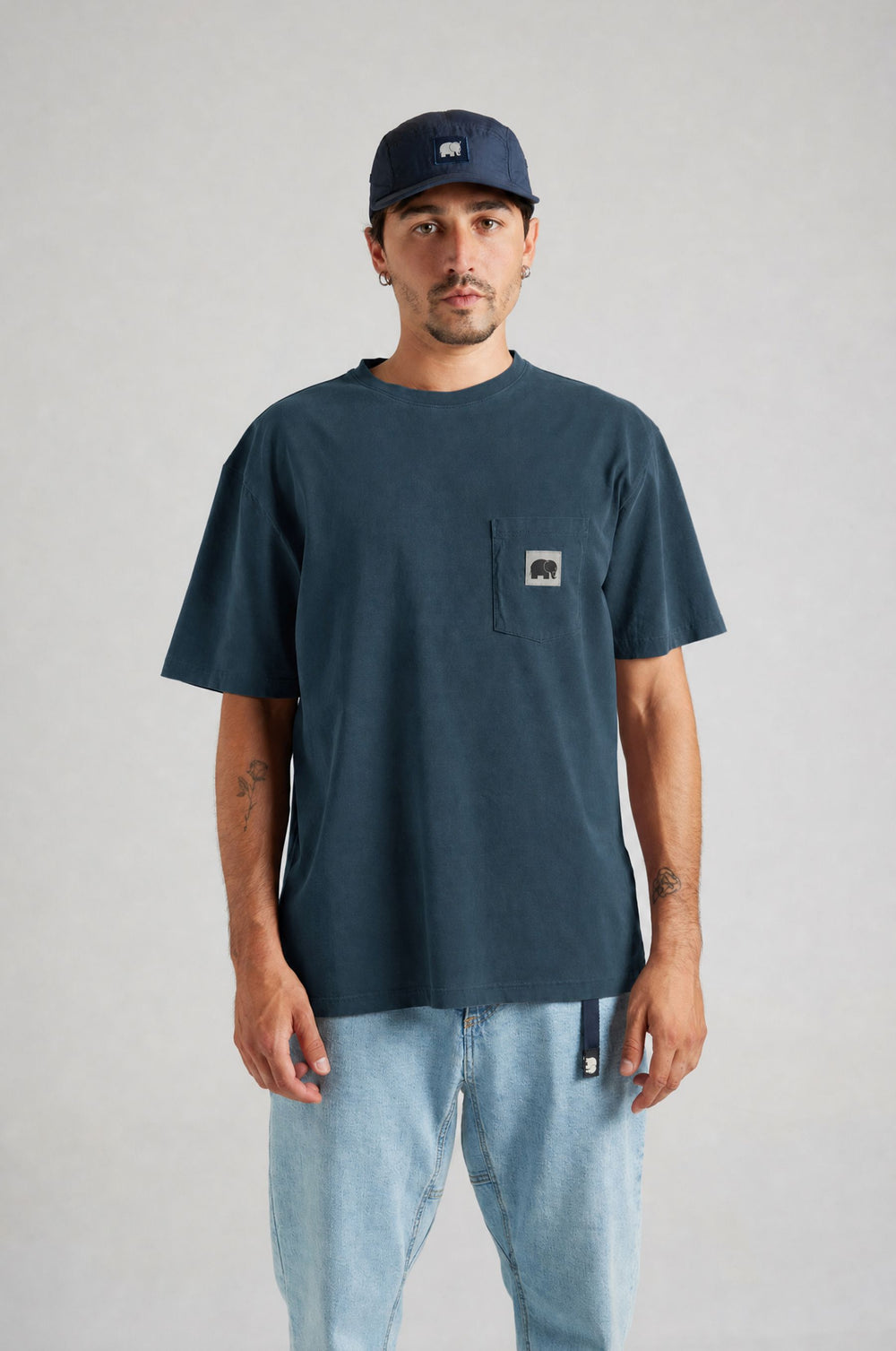 Persona che indossa una t-shirt blu scuro con una tasca e un logo a forma di elefante, abbinata a jeans azzurri | TP_IMAGE_TYPE=MODEL_FRONT