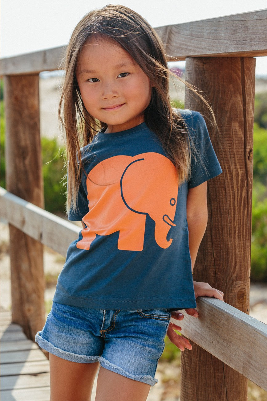 Personne portant un Kid's Organic Classic T-Shirt en Trendsplant Blue avec un motif d'éléphant orange, posant en extérieur sur une terrasse en bois. | TP_IMAGE_TYPE=MODEL_FRONT