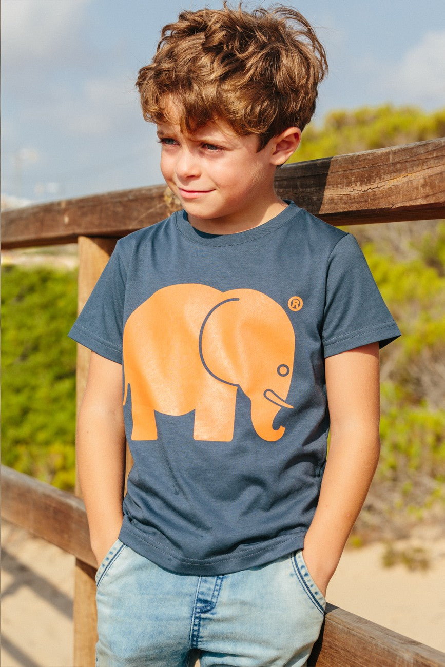 Personne portant un Kid's Organic Classic T-Shirt bleu avec un graphique d'éléphant orange, souriant debout près d'une rambarde en bois. | TP_IMAGE_TYPE=MODEL_FRONT