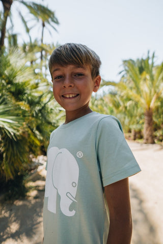 Persona che indossa una t-shirt classica biologica verde menta con una grafica raffigurante un elefante, sorridente in un ambiente esterno soleggiato circondata da palme. | TP_IMAGE_TYPE=MODEL_FRONT