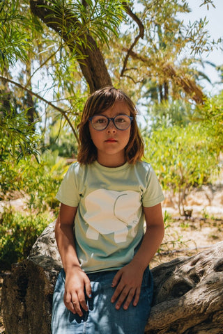Persona che indossa una t-shirt classica biologica verde menta con il disegno di un elefante, seduta su un tronco tra la vegetazione. | TP_IMAGE_TYPE=MODEL_FRONT