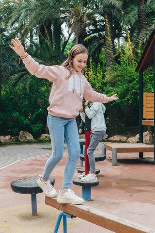 Persona che indossa una Kid's Organic Classic Hoodie in Pale Pink mentre mantiene l'equilibrio su una trave del parco giochi | TP_IMAGE_TYPE=LIFESTYLE