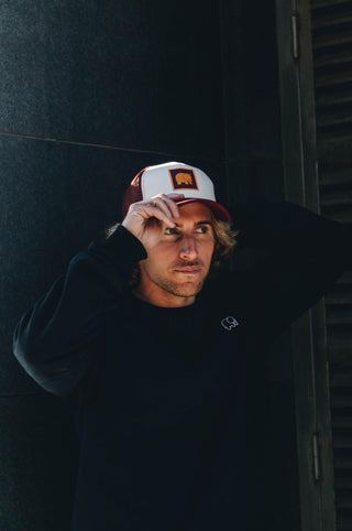 Personne portant une casquette trucker classique bordeaux, en train de l'ajuster tout en regardant pensivement hors caméra | TP_IMAGE_TYPE=MODEL_FRONT