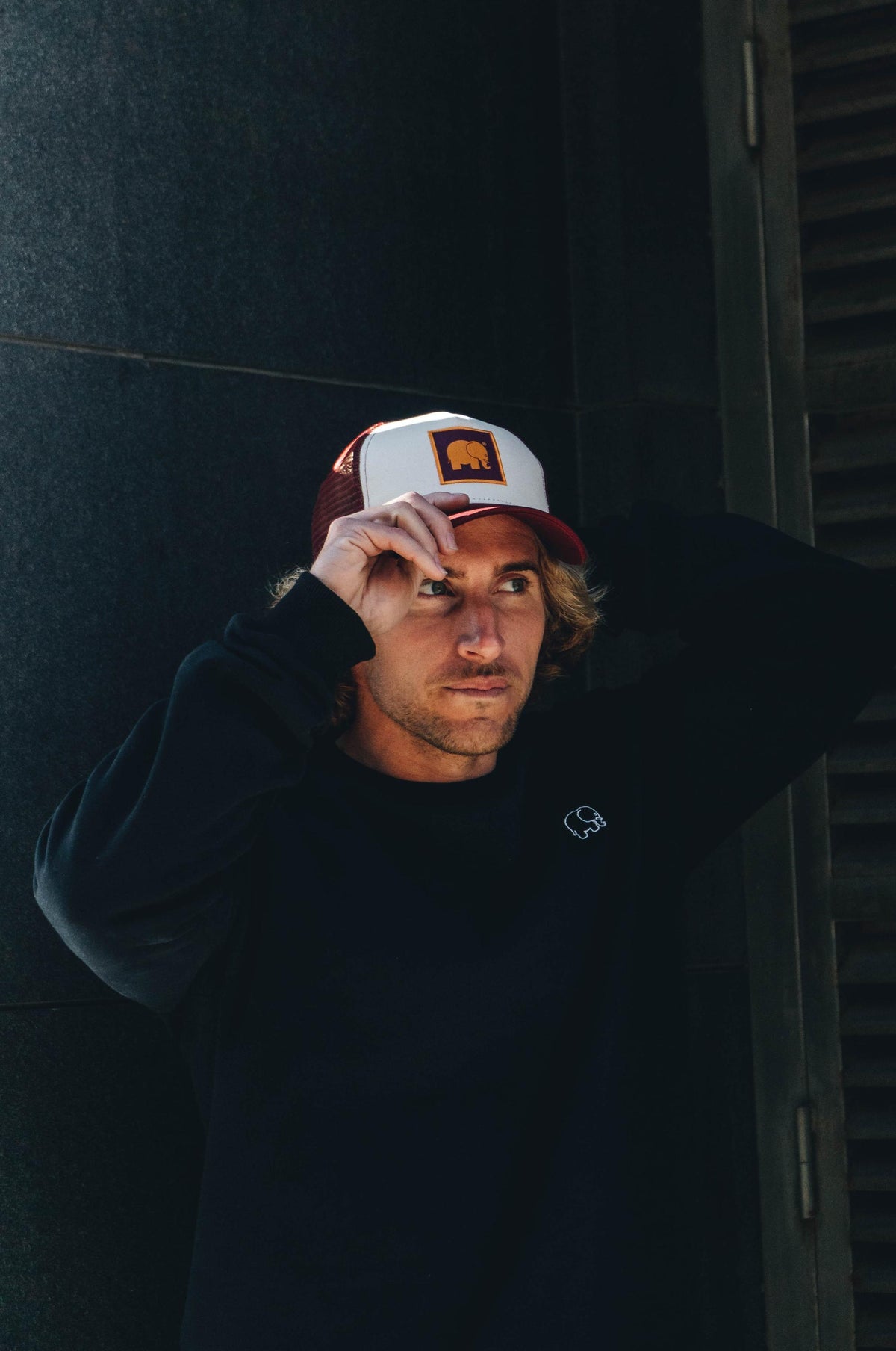 Personne portant une casquette trucker classique bordeaux, en train de l'ajuster tout en regardant pensivement hors caméra | TP_IMAGE_TYPE=MODEL_FRONT